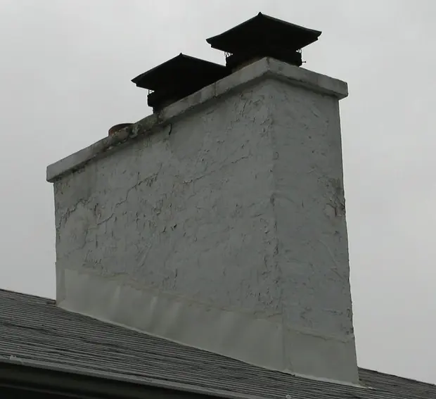Wide painted masonry chimney with two black chimney covers on a shingled roof