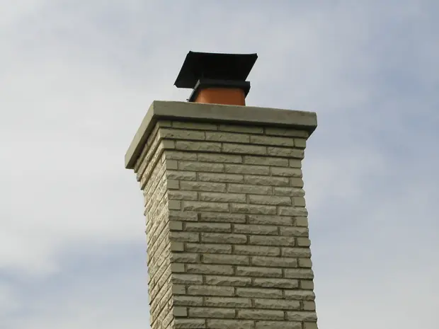 Rebuilt light brick chimney with a projecting concrete crown and a black chimney cover against a blue sky