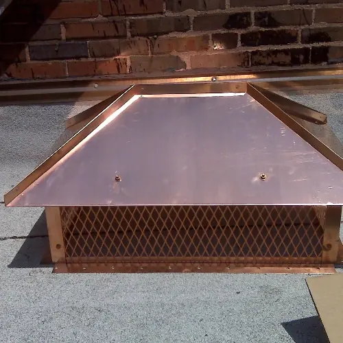 Newly fabricated copper hip-and-ridge chimney cover with mesh sides, sitting on a roof