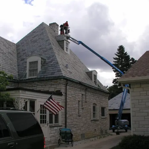 Boom lift reaching over a slate roof to the chimney of a stone house
